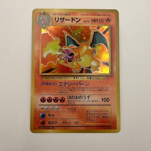 Puede incluir: Una carta coleccionable japonesa de Charizard con una ilustración holográfica de la criatura naranja parecida a un dragón escupiendo fuego. La carta muestra el nombre リザードン, HP120 y varias estadísticas de juego, incluyendo poder de ataque y requisitos de energía impresos en texto japonés sobre un fondo naranja ardiente.