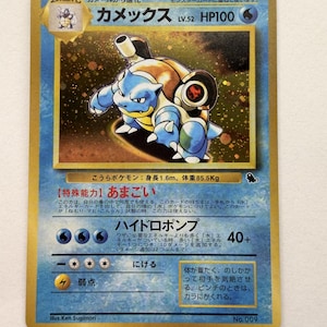 May include: A Japanese Blastoise trading card with a gold border, featuring the blue water-type Pokémon with cannons on its back. The card displays HP100, the name Kamex, and Japanese text detailing its special ability and Hydro Pump attack.