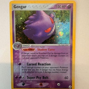 May include: A Gengar holographic trading card with a purple character illustration, 100 HP, and the attacks Shadow Curse, Cursed Reaction, and Super Psy Bolt. The card features a yellow border, a stage 2 evolution label, and 2006 copyright text.