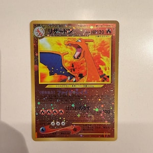 May include: A Japanese Charizard Pokémon trading card with a gold holographic finish. The card features an illustration of the fire-type dragon, HP120, and Japanese text including the name Lizardon. The background has a star-patterned foil effect.