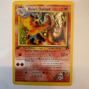 Puede incluir: Una carta coleccionable de Blaine's Charizard con una ilustración holográfica del Pokémon de tipo fuego. La carta muestra el nombre, 100 HP, evolución de fase 2, los ataques Roaring Flames y Flame Jet, y un sello de 1ª edición en borde amarillo.