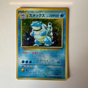 May include: A Japanese Blastoise trading card featuring a holographic illustration of the blue turtle-like creature. The card displays the name Kamex, HP100, and water-type symbols. Text includes special ability Amagoi and Hydro Pump attack details.