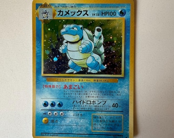 Può includere: Una carta collezionabile giapponese di Blastoise con un'illustrazione olografica della creatura blu simile a una tartaruga. La carta mostra il nome Kamex, 100 HP e simboli di tipo acqua. Il testo include la capacità speciale Amagoi.