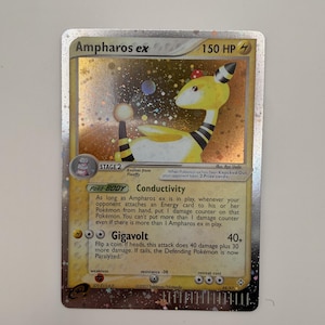 May include: A holographic Ampharos ex trading card with 150 HP, featuring a yellow and black creature illustration. The card displays the text Conductivity and Gigavolt, along with game mechanics, set information, and a sparkling foil finish.