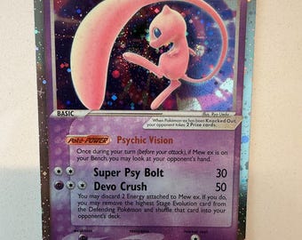 Può includere: Una carta collezionabile Mew ex con uno sfondo olografico viola e un'illustrazione del personaggio Mew rosa. La carta mostra 90 HP, il Poke-Power Psychic Vision e gli attacchi Super Psy Bolt e Devo Crush, con testo sulle regole di gioco.