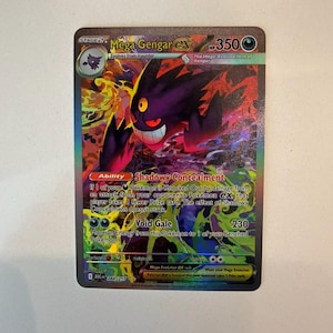 Puede incluir: Una carta coleccionable Mega Gengar EX con 350 HP, que muestra una criatura de color púrpura oscuro con un ojo amarillo. La carta presenta la habilidad Shadowy Concealment y el ataque Void Gale sobre un fondo vibrante y arremolinado en verde, amarillo y rojo.
