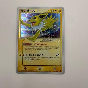 Puede incluir: Una carta coleccionable Pokémon Jolteon Gold Star japonesa que muestra una criatura eléctrica amarilla con un cuello blanco puntiagudo. La carta tiene un fondo holográfico con patrones de rayos, 70 HP y texto en japonés.