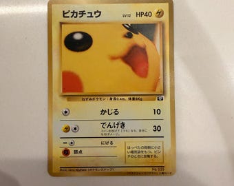 Può includere: Una carta collezionabile di Pikachu con una fotografia in primo piano del volto del personaggio. La carta riporta il nome Pikachu, livello 12, 40 HP e testo in giapponese che include nomi e descrizioni degli attacchi. Bordo dorato.