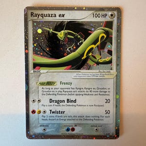 May include: A holographic Rayquaza ex trading card with 100 HP, featuring a green serpentine creature against a dark swirling background. The card displays the attacks Dragon Bind and Twister, along with game text, energy symbols, and a 2006 copyright.