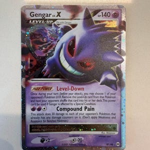May include: A Gengar LV.X trading card with a purple holographic finish. The card features the character Gengar, 140 HP, the Level-Down Poke-Power, and the Compound Pain attack. It includes the text 97/99 and 2009 Pokemon/Nintendo at the bottom.