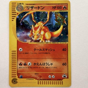 May include: A Japanese Charizard trading card with a gold border, featuring a holographic illustration of the orange dragon-like creature with blue wings against a starry background, displaying 100 HP and two fire-type attack moves.