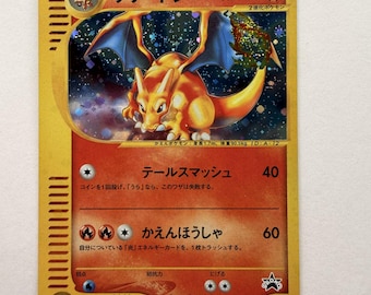Può includere: Una carta collezionabile giapponese di Charizard con bordo dorato, caratterizzata da un'illustrazione olografica della creatura arancione con ali blu su sfondo stellato, con 100 HP e due attacchi di tipo fuoco.