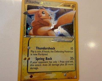 Può includere: Una carta collezionabile gialla Pikachu Gold Star con un'illustrazione olografica del personaggio con le braccia incrociate. La carta mostra 60 HP, gli attacchi Thundershock e Spring Back e il testo 2006 Pokemon Nintendo in basso.