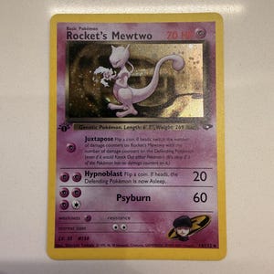 Può includere: Una carta collezionabile Rocket's Mewtwo con bordo viola e cornice gialla. La carta presenta il personaggio Mewtwo, 70 HP e gli attacchi Juxtapose, Hypnoblast e Psyburn. Include il logo del Team Rocket, il timbro 1st Edition e del testo.
