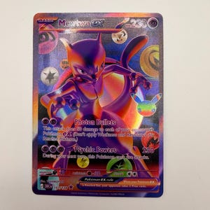 Puede incluir: Una carta coleccionable Mewtwo ex con un diseño holográfico en morado y naranja. La carta muestra al personaje Mewtwo, el texto Photon Bullets, Psychic Powers y 230 HP. Incluye varios símbolos de energía y el número de carta 157/128.