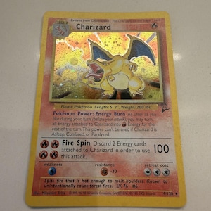 May include: A holographic Charizard trading card featuring a yellow border, a fire-type dragon illustration, and text detailing the Energy Burn power and Fire Spin attack. The card displays 120 HP, level 76, and the number 4/130 at the bottom.