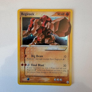 May include: A Regirock trading card with 90 HP, featuring a brown and red rock-like creature illustration. The card text includes Dig Drain and Final Blast attacks, with a yellow border and a holographic background behind the character artwork.