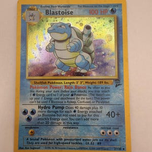 Peut inclure: Une carte à collectionner holographique Blastoise avec une bordure jaune. La carte affiche le personnage, 100 PV, le pouvoir Pokémon Danse Pluie et l'attaque Hydrocanon. Elle indique une taille de 1,6 m et un poids de 85,7 kg. Niveau 52.