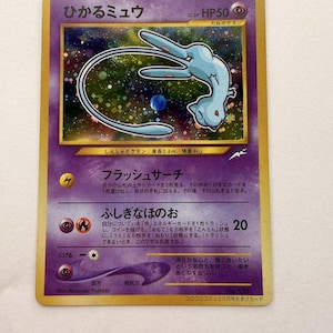 Puede incluir: Una carta coleccionable Pokémon Shining Mew japonesa con borde morado y detalles en lámina dorada. La carta presenta una ilustración holográfica del personaje Mew azul claro, HP50, y texto en japonés, incluido el nombre Hikaru Mew.
