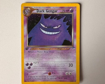 Può includere: Una carta collezionabile Dark Gengar con un'illustrazione di un personaggio viola simile a un fantasma, 70 HP e il testo 'Shadow Pokémon. Length: 1.5 m, Weight: 40.4 kg.' La carta presenta un bordo giallo, simboli di energia psichica.