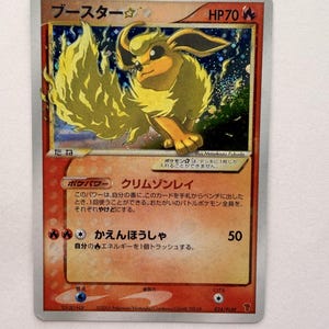 Puede incluir: Una carta coleccionable japonesa Flareon Gold Star con una ilustración holográfica de la criatura de tipo fuego con una cola grande y esponjosa. La carta muestra el nombre en japonés, 70 HP, texto del juego, ataques y copyright de 2005.