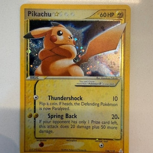 May include: A yellow Pikachu trading card featuring a holographic illustration of the character with crossed arms. The card displays 60 HP, the attacks Thundershock and Spring Back, and text stating you cannot have more than one Pokemon star in your deck.