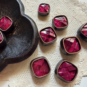 Op de afbeelding: Een verzameling vierkante knopen met een diep magenta, gefacetteerd middenstuk omringd door een donker, metaalkleurig frame. De knopen hebben een geometrische, veelzijdige oppervlaktestructuur die het licht reflecteert voor een glanzend effect.