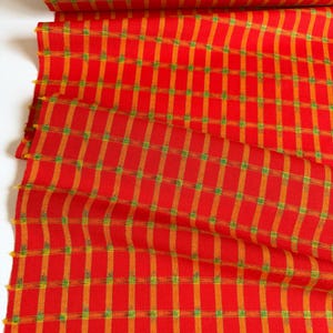 May include: A bolt of fabric featuring a vibrant red base color with a repeating grid pattern. The design consists of vertical and horizontal orange stripes intersected by small, bright green squares at each crossing point of the plaid motif.