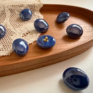May include: A collection of navy blue, oval-shaped ceramic buttons with a glossy finish, displayed on a wooden tray. One button has a gold-colored shank. The buttons are arranged on a white lace doily.