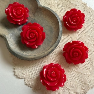Puede incluir: Cinco adornos brillantes en forma de rosa de color rojo intenso con pétalos en capas. Dos de los adornos de rosa están colocados dentro de un cuenco de madera gris de forma ovalada con un interior tallado liso, mientras que tres están sobre un tapete de encaje color crema.