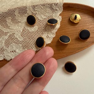 以下が含まれることがあります： 金色の縁取りが付いた黒い丸いボタンのコレクション。ボタンは木製のトレイとレースの上に配置されています。ボタンの質感とデザインを示すために、1つのボタンが手に持たれています。