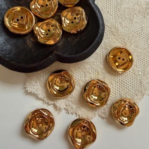 May include: A collection of shiny gold-toned buttons with a smooth, reflective finish. Some buttons feature a circular design with a central square indentation, while others have a wavy, flower-like edge. Each button has two small holes.