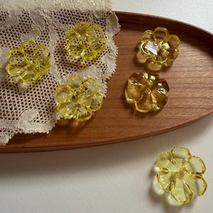 以下が含まれることがあります： 黄色い花の形をした6つの装飾品。花は半透明の素材で作られており、詳細な花びらのデザインが施されています。木製のトレイとレースのドイリーに配置されています。花は明るく日当たりの良い色をしています。
