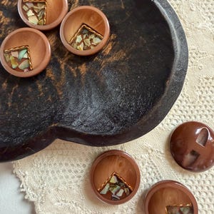 以下が含まれることがあります： 中央が窪んだ5つの明るい茶色のボタン。窪みには虹色に輝く多色の貝殻の破片が三角形にはめ込まれ、細い金色の金属ワイヤーで縁取られています。また、2つの長方形の穴が開いた濃い茶色のボタンが1つあります。