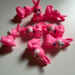 May include: A collection of seven small, bright pink plastic rabbit figurines. Each rabbit features white painted facial details, large dark eyes, and a small blue bowtie. The figures are arranged in a scattered pattern, showing various poses including sitting upright and lying down on their sides.