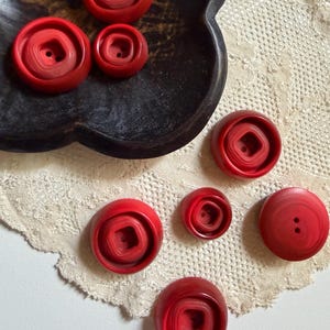 以下が含まれることがあります： 7つの赤い丸いボタンのコレクション。それぞれ中央が四角くくぼんでおり、2つの穴が開いています。6つのボタンは同心円状の段差があるデザインで、1つは平らで2つの穴が開いています。ボタンのサイズはそれぞれ異なります。