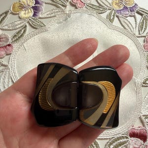 以下が含まれることがあります： 光沢のある黒いベースに、抽象的なゴールドトーンの金属アクセントと滑らかな曲線を描くベージュのラインが施された、長方形のヘアクリップのペア。クリップは磨き上げられた仕上げで、質感のあるゴールドのインサートを備えた左右対称のデザインです。