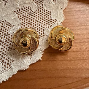 以下が含まれることがあります： 花のような円形デザインのゴールドトーンの金属製イヤリングのペア。各イヤリングには、滑らかで光沢のある中央のドームがあり、その周囲を質感のある渦巻く花びらが囲んでおり、金属仕上げで層状の立体的な外観を作り出しています。