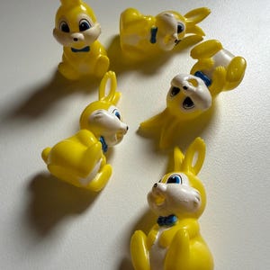 以下が含まれることがあります： 顔と胸に白いアクセントがある、5体の小さな黄色いプラスチック製のウサギのフィギュア。各ウサギには青く塗られた目と、首の周りに小さな青い蝶ネクタイが付いています。フィギュアはさまざまな座った姿勢や横たわった姿勢で配置されています。