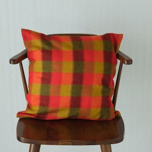 May include: A square throw pillow featuring a vibrant plaid pattern in shades of mustard yellow, bright orange, and deep maroon. The pillow is positioned on a dark brown wooden chair with curved armrests and a solid backrest, set against a plain wall.