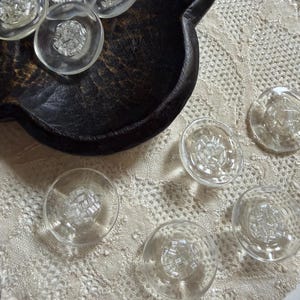 May include: A collection of round, clear glass buttons featuring a textured, grid-like pattern in the center. The buttons have a smooth, transparent outer rim and are arranged both inside a dark, carved wooden bowl and on a lace fabric surface.