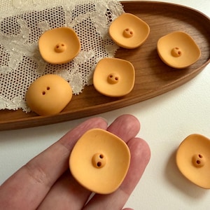 Op de afbeelding: Een verzameling van zeven gladde, matte, perzikkleurige knopen met een licht gebogen, vierkante vorm. Elke knoop heeft twee centrale gaten voor bevestiging. De knopen hebben een egale kleur en een ontwerp met zachte, afgeronde randen.