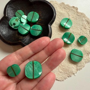 May include: A collection of round, bright green buttons with a pearlescent finish and a single dark vertical line running through the center. These buttons vary in size and feature a smooth, polished surface with a shank on the back for attachment.