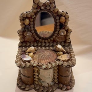 May include: A decorative vanity mirror and storage unit, intricately adorned with various seashells. The mirror is oval-shaped, and the unit features two small drawers and a central shelf, all embellished with a variety of shells.