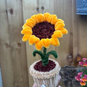 Pode incluir: Um girassol artesanal feito de limpadores de cachimbo felpudos, com pétalas amarelo-brilhantes e um centro castanho-escuro. A flor está num pequeno vaso bege texturizado preenchido com material castanho-escuro, sobre um caule verde.