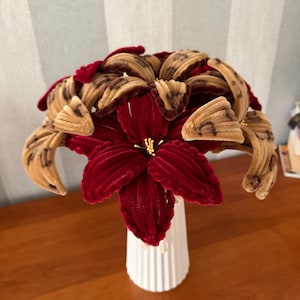 May include: A bouquet of handcrafted flowers made from fuzzy pipe cleaners in deep red and tan with dark brown leopard print spots. The flowers feature prominent petals and thin yellow stamens, arranged in a white, vertically ribbed cylindrical vase.