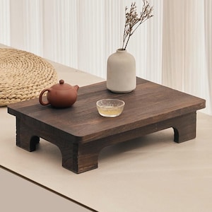 May include: A dark brown, low wooden table with a rectangular top and four short legs. A small brown teapot, a clear glass bowl, and a white vase with dried stems are on the table. The table is likely used for serving tea.