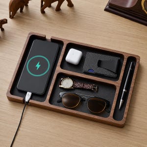 以下が含まれることがあります： 充電アイコンが表示されたスマートフォン、白いワイヤレスイヤホン、カーボンファイバー製財布、茶色のレザーベルトの腕時計、サングラス、黒いペンが入った、複数の仕切りがある長方形の木製トレイ。