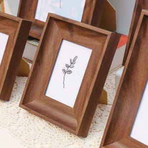May include: A set of rectangular wooden picture frames with a dark brown finish and a smooth, angled border. Each frame contains a white insert featuring a simple black line drawing of a single leafy plant stem centered on the white background.