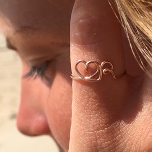Könnte beinhalten: Ein zarter Ring aus Roségold mit einem Herz- und Wellenmuster. Der Ring wird an einem Finger einer Person mit blonden Haaren getragen.